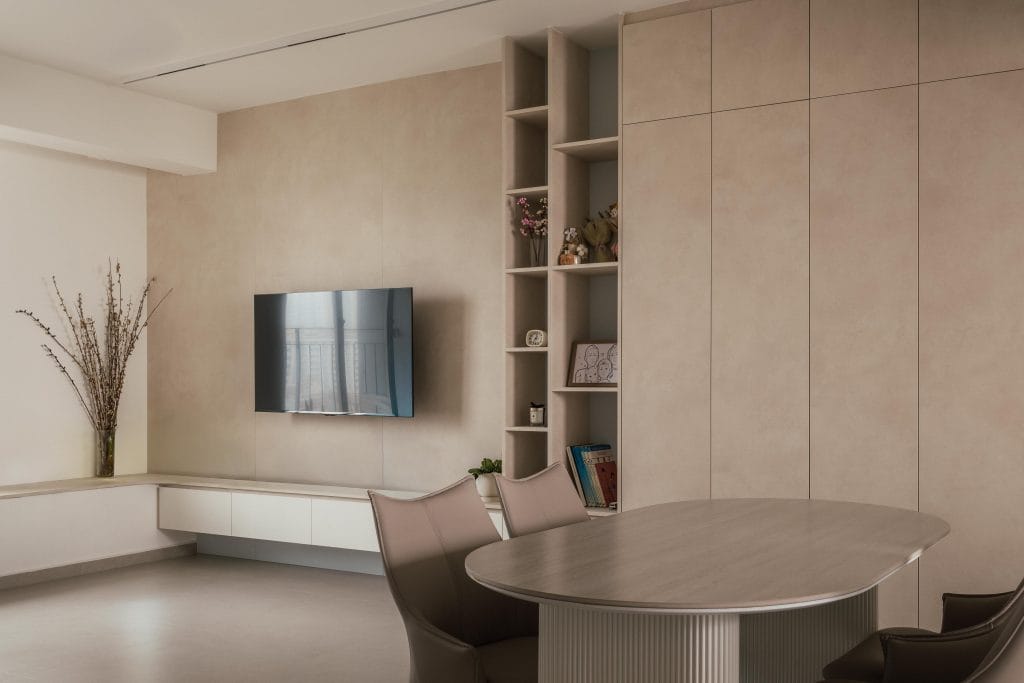 A warm minimalist living and dining space at 78 Mergui Road featuring a curved dining table with a fluted base, brown leather chairs, seamless beige built-in cabinets, and a wall-mounted TV.