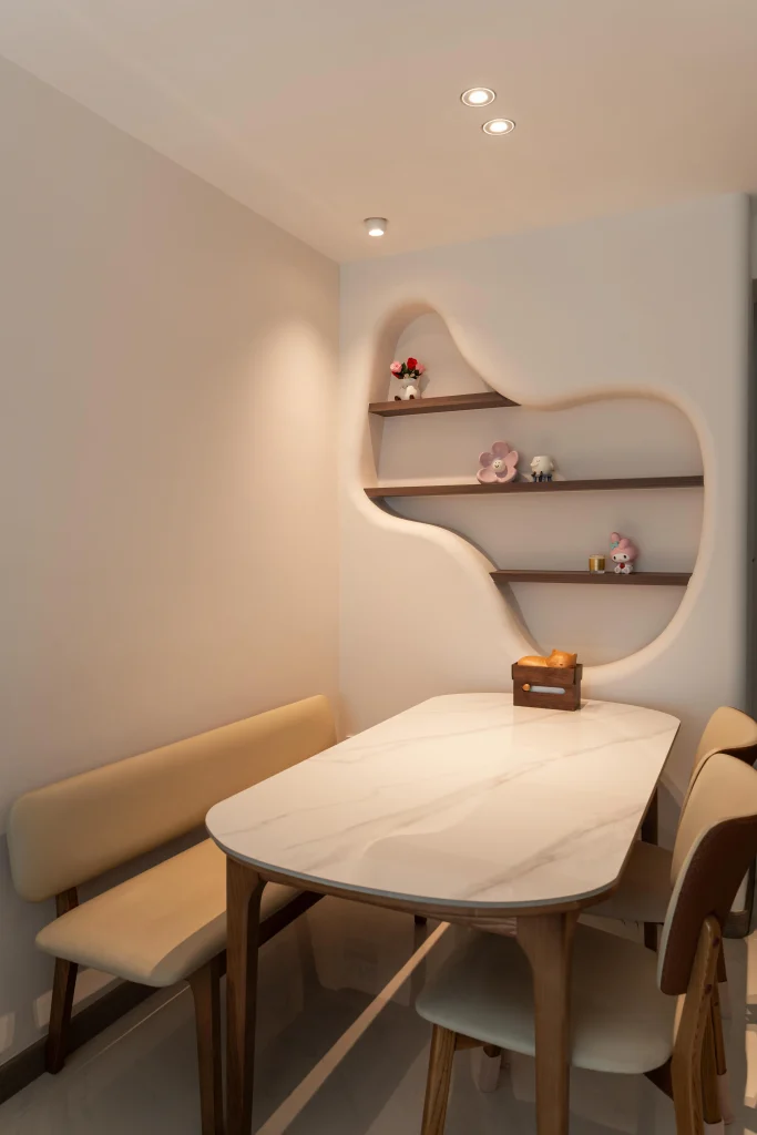 A modern dining area at 322A Tengah Drive featuring a white marble-topped table, a beige upholstered bench, and an organically shaped illuminated wall niche with wooden display shelves.