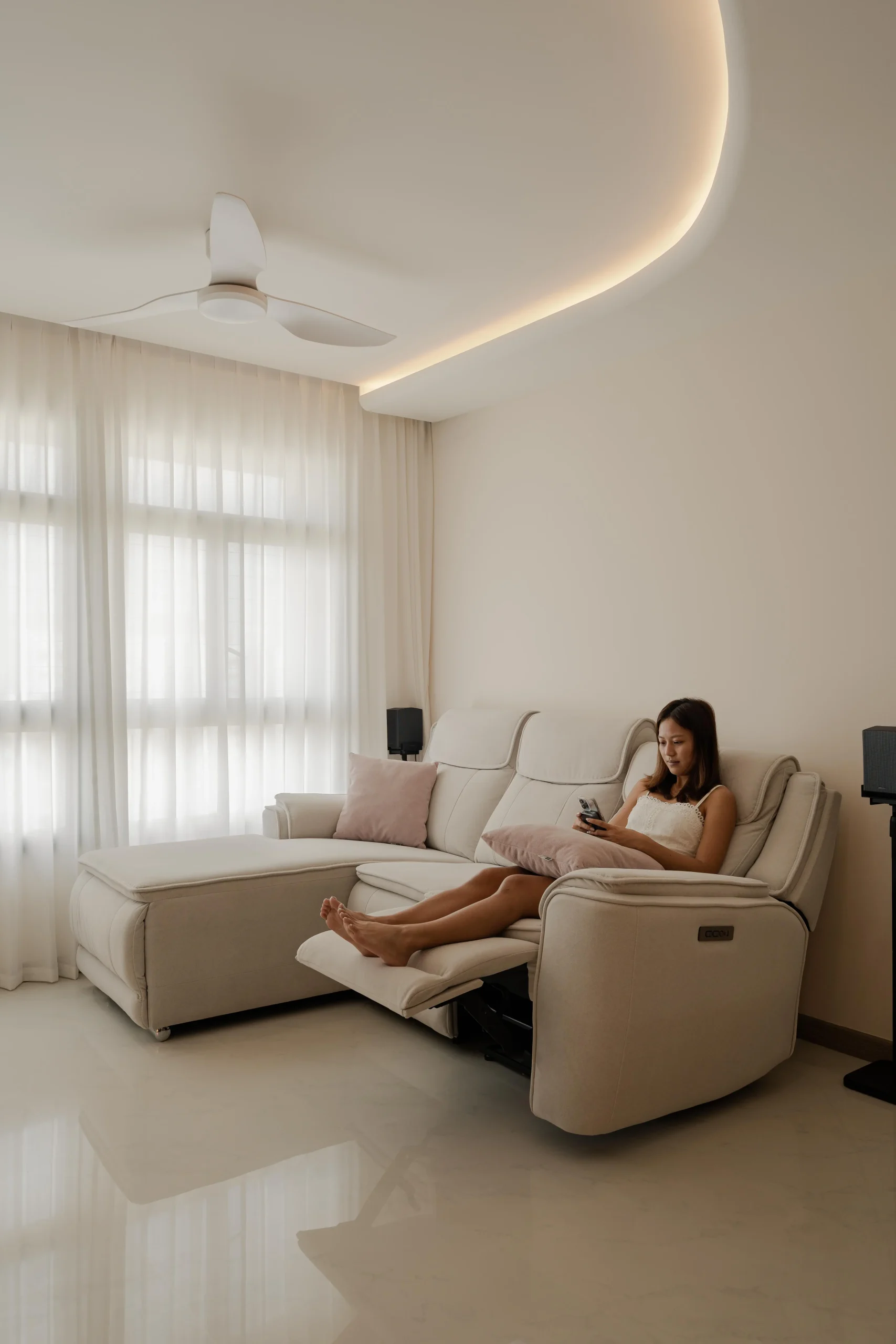 A woman relaxing and looking at her phone on a light-colored reclining sectional sofa in a bright living room at 322A Tengah Drive, featuring sheer white curtains, a ceiling fan, and curved cove lighting.